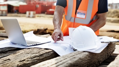 close up shot of a table with building plans being inspected bu a person in an orange high-vis vest. theer's a white hard hat on the table.