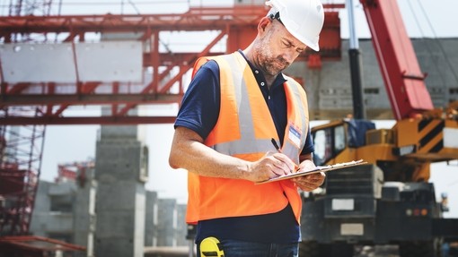 man in an orange hi-vis vest and white hard hat looking at plans. there is a construction site in the background