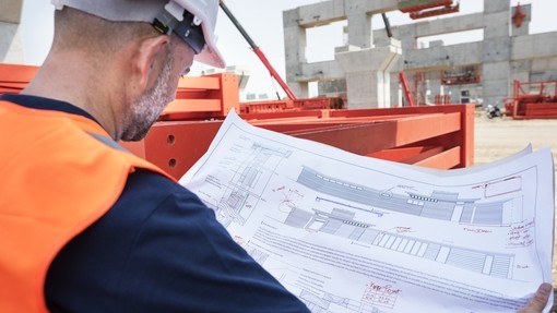 over the shoulder shot of a man in orange high-vis vest and white hard hat looking at building plans