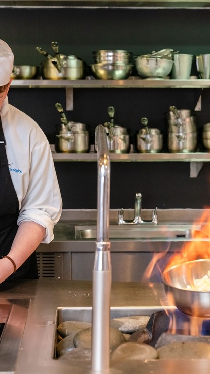 Two students wearing chef whites and aprons. One student is flambéing.