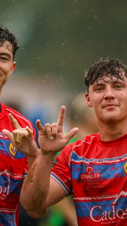 Two students in muddy rugby kit smiling at the camera and making positive hand gestures.