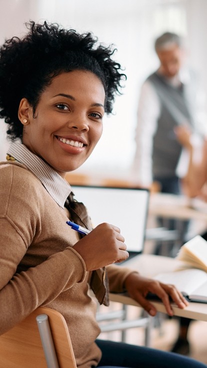 black female learner smiling at the camera, behind her the tutor provides support to other learners