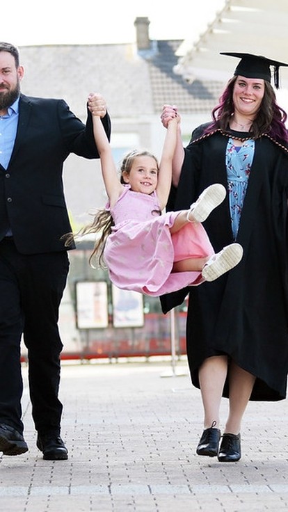 a woman in a graduation cap and gown, she and her partner are swinging their daughter in the air between them.