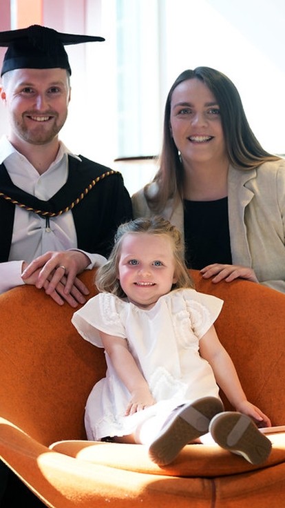 A man in a graduation cap. he and his wife stand behind a round orange chair where their young daughter is sitting.
