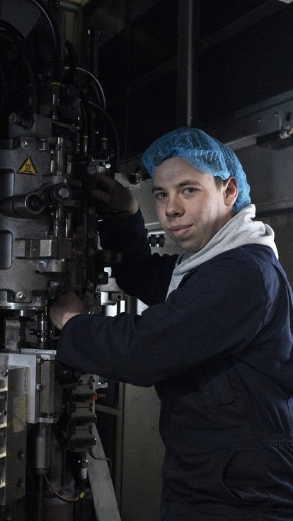 male apprentice in dark blue overalls and a hairnet in front of industrial machinery