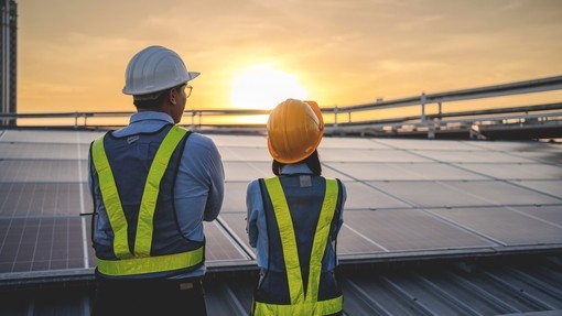 Two managers, one male, one female. Both wearing hivis and har hats while looking towards solar panels, their back turned to the camera