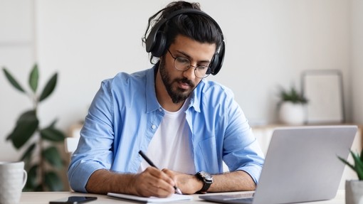 A male wearing headphones makes notes while on his laptop at home