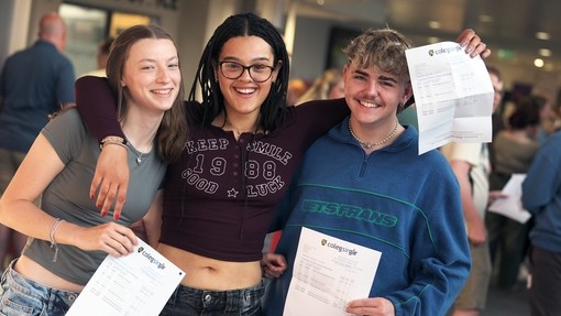 Three students stood together smiling and showing their exam results.