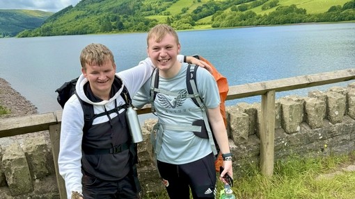 Two students stopping for a photo, one with their arm around the other with mountains and water in the background