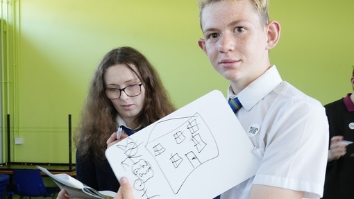 Two pupils taking part in a Welsh workshop with one looking at the camera holding up some work