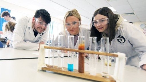 A lecturer and two students in white lab coats and goggles, looking at a chemical reaction