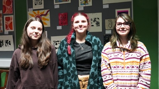 Three students in a row with their work displayed on a wall behind them