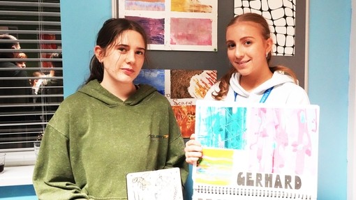 Two students holding up work and looking at the camera with their work on the wall behind them