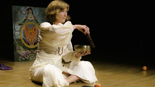 Milly, sitting on a performance type floor wearing all white using a singing bowl