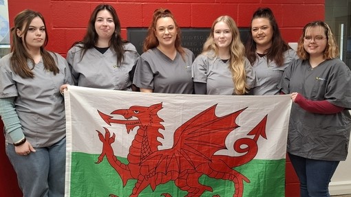 Six students standing in line with a red background holding a Welsh flag
