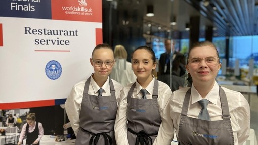 Three students in grey/smart aprons and ties in front of a restaurant service final sign