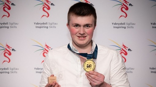 Twm (medallist) in a shirt and tie holding up his medal with Inspiring Skills branding behind him