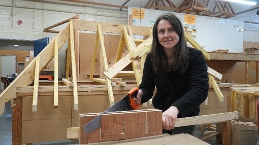 Jess looking at the camera with a saw in her hand at a workbench with a timber frame in the background