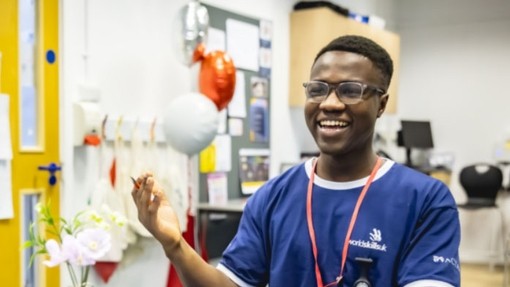A young person in a blue t-shirt branded WorldSkillsUK at a desk wearing a lanyard smiling