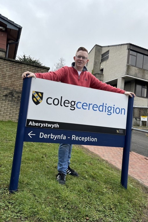 Cerith in his red ambassador hoodie standing behind the Coleg Ceredigion sign