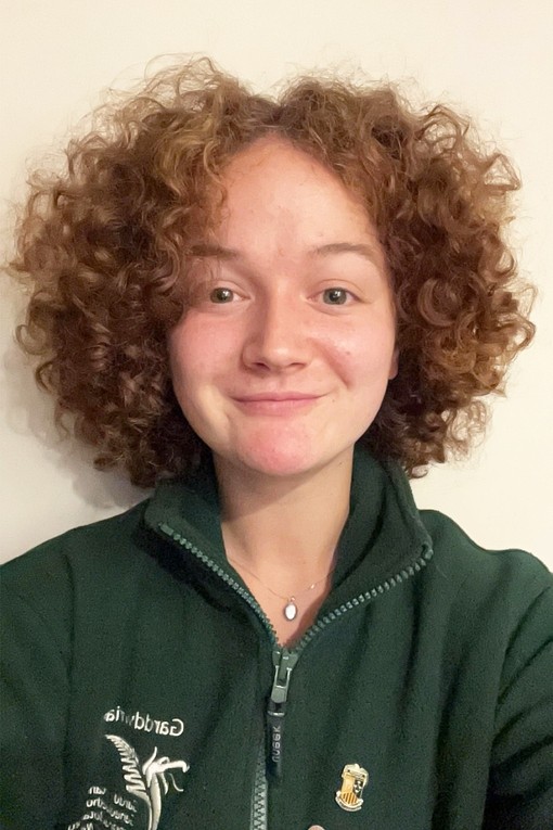 Katie, who has shoulder length curly hair, who is looking at the camera in a green National Botanic Gardens fleece