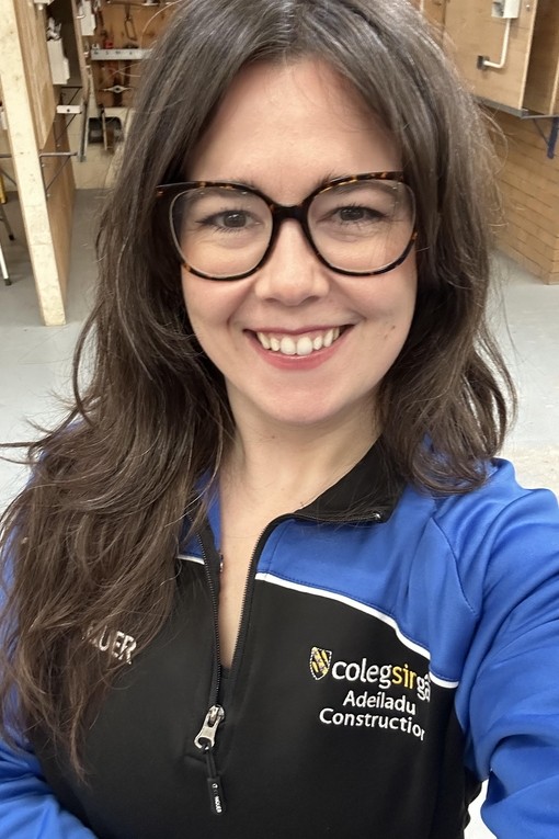 Abigail in the carpentry workshop wearing a college branded blue and black top looking at the camera smiling wearing glasses