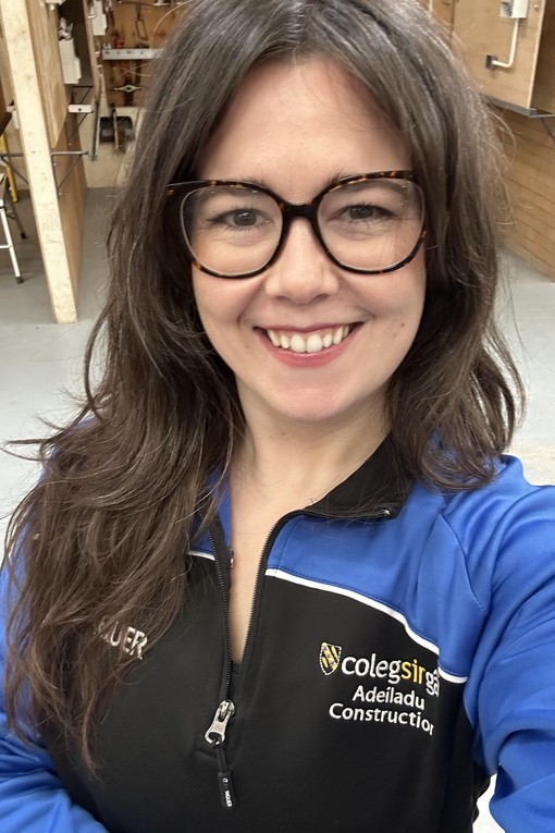 Abigail in the carpentry workshop wearing a college branded blue and black top looking at the camera smiling wearing glasses