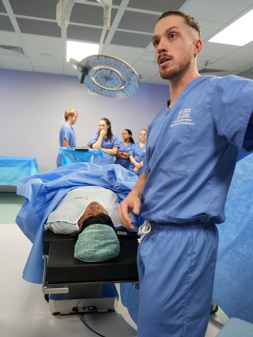 A man in blue scrubs with a theatre bed behind talking to students