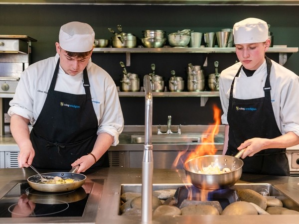 Two students wearing chef whites and aprons. One student is flambéing.