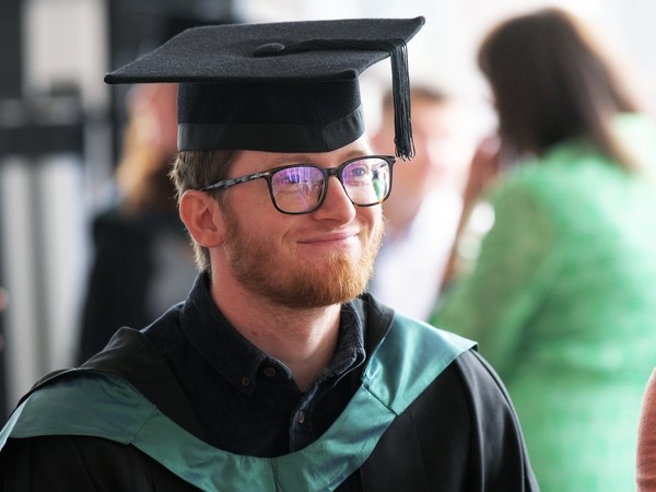 Smiling student with glasses and a beard, wearing a graduation cap and gown.
