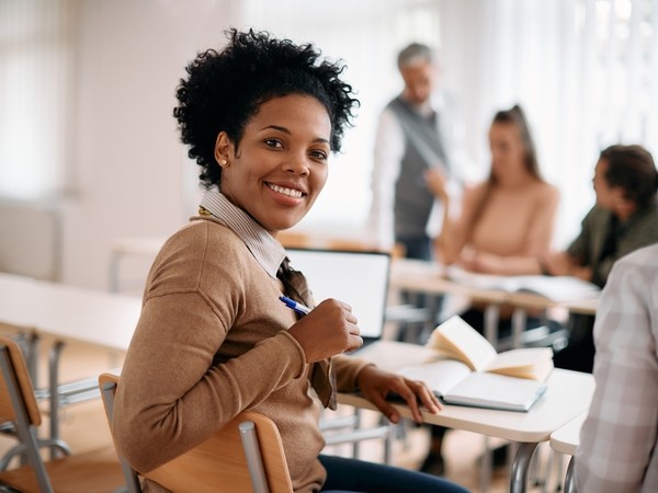 black female learner smiling at the camera, behind her the tutor provides support to other learners