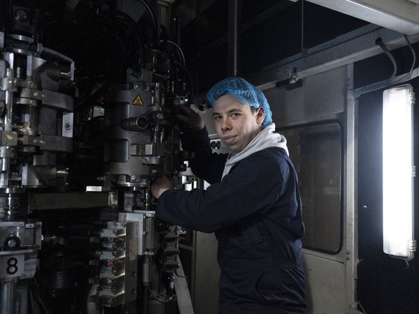 male apprentice in dark blue overalls and a hairnet in front of industrial machinery