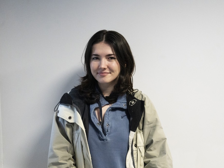 Olivia standing against a plain wall looking at the camera in a blue top and coat