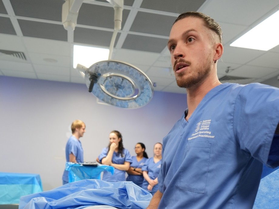 A man in blue scrubs with a theatre bed behind talking to students