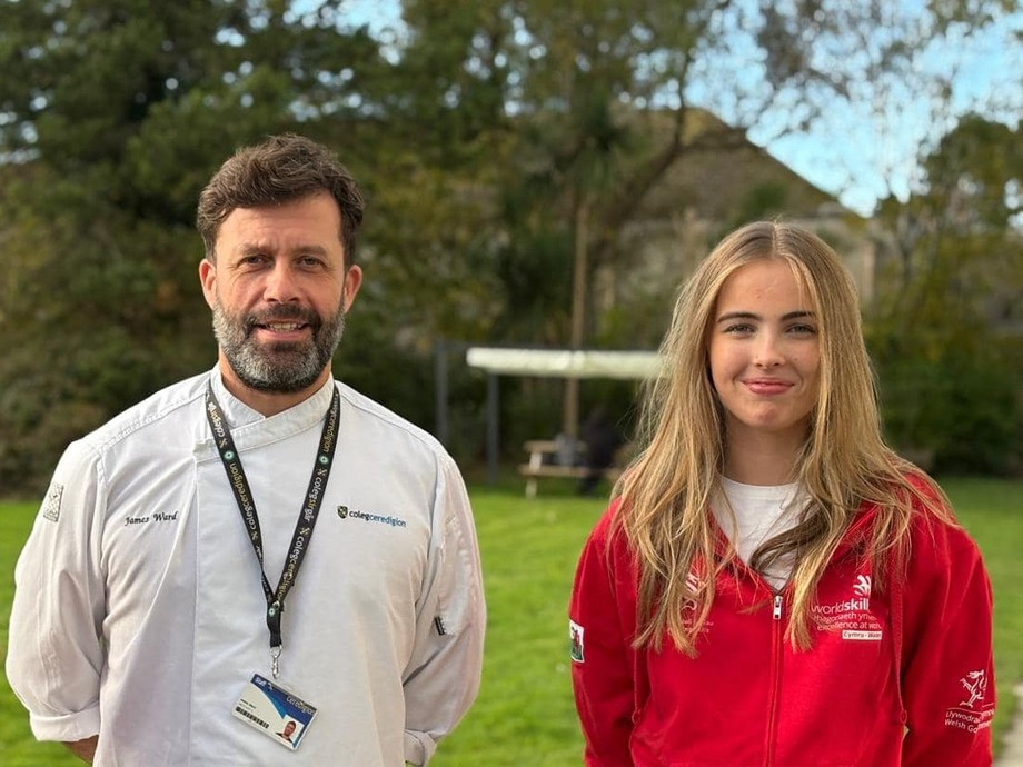 Caitlin in a red Wales top standing next to James Ward lecturer in chef whites with grass in the background