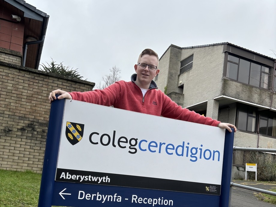 Cerith in his red ambassador hoodie standing behind the Coleg Ceredigion sign