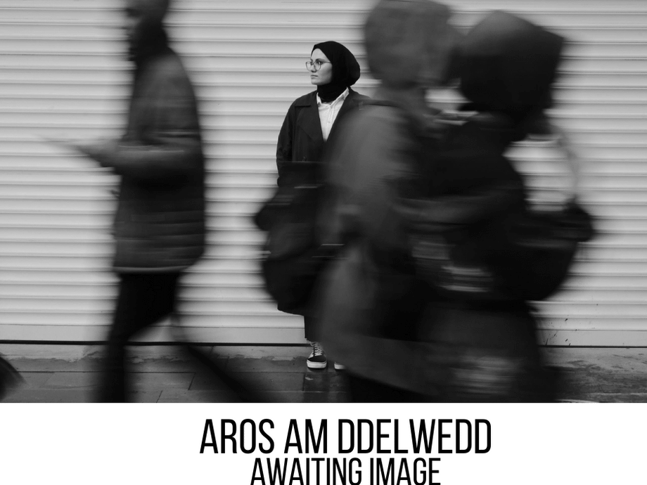 An image of a woman lent against a shutter with blurred people walking past it is black and white and a creative shot
