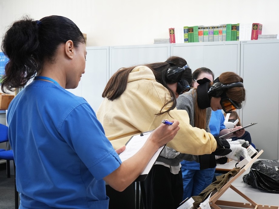 A nurse in the foreground writing something on a clipboard with students working in the background