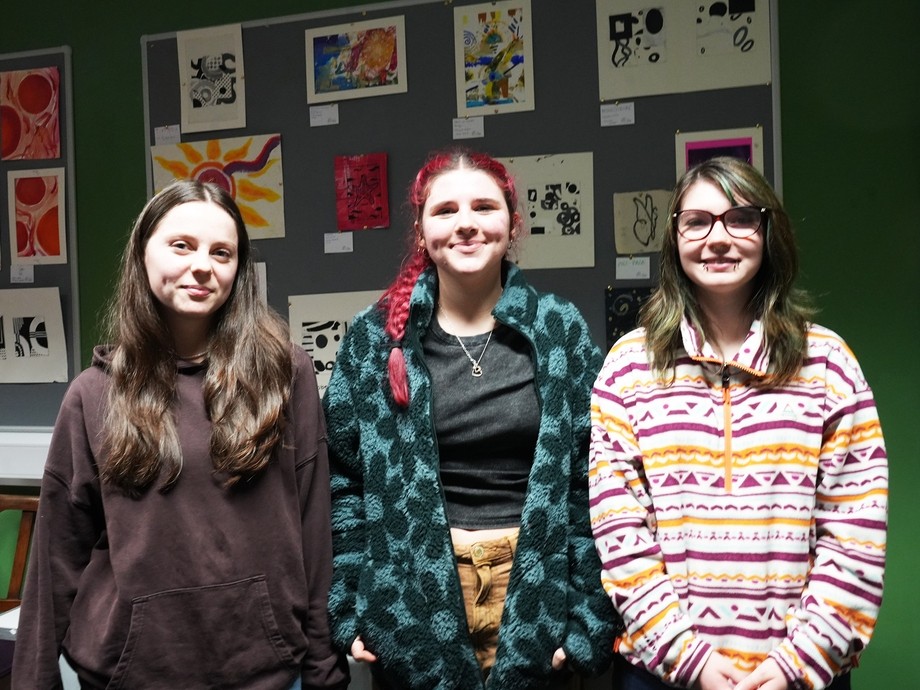 Three students in a row with their work displayed on a wall behind them