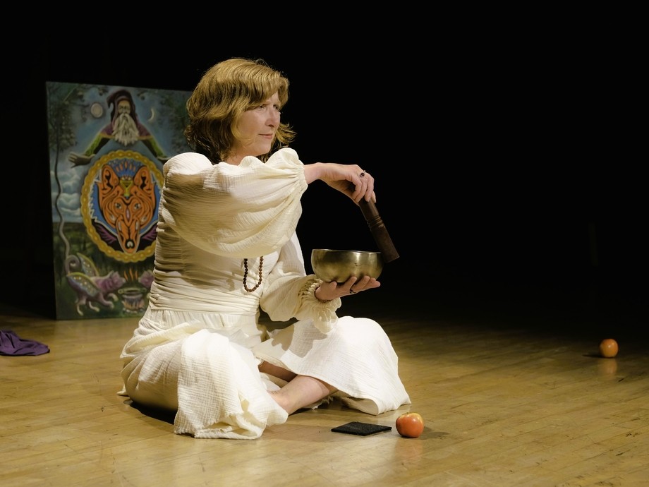 Milly, sitting on a performance type floor wearing all white using a singing bowl