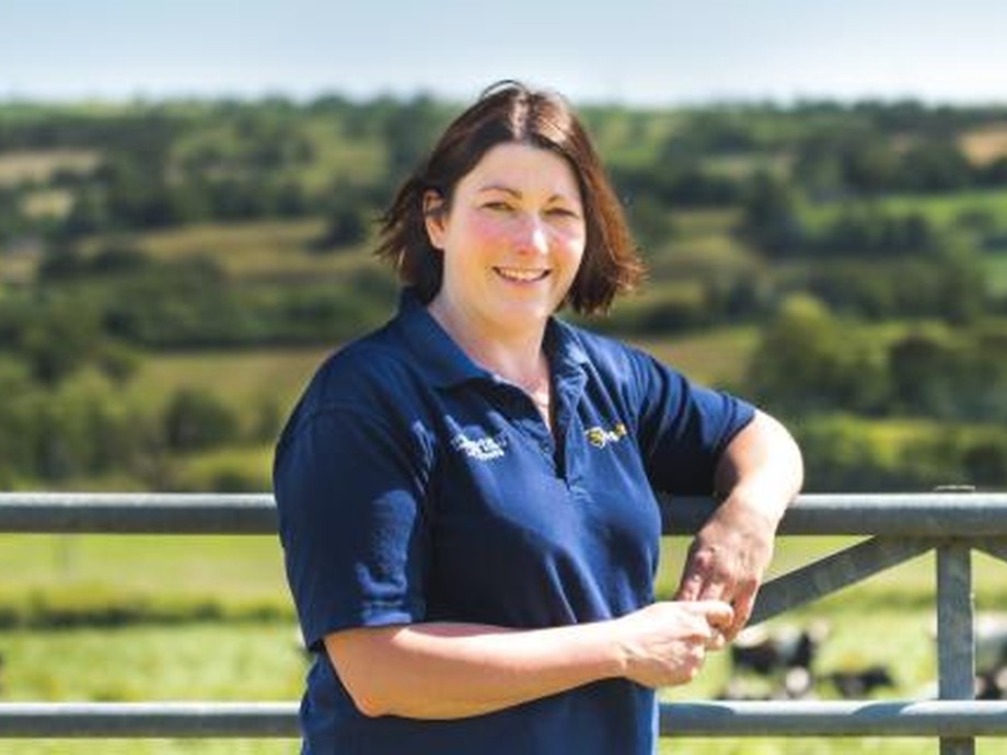 Liz standing and leaning on a fence outside with grassland behind her looking at the camera