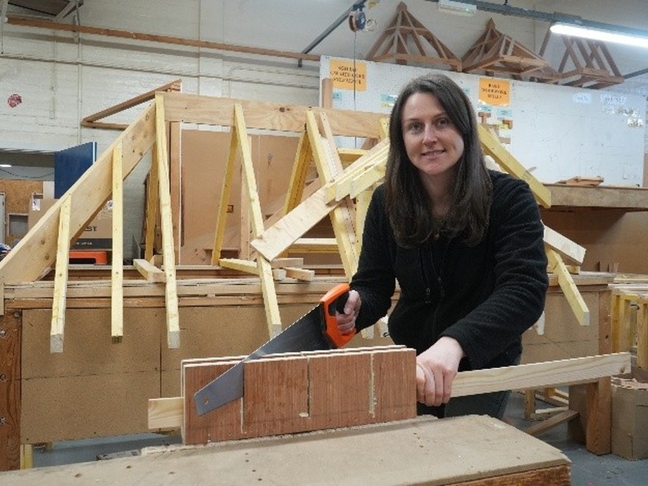 Jess looking at the camera with a saw in her hand at a workbench with a timber frame in the background