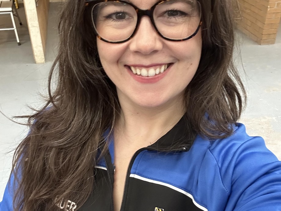 Abigail in the carpentry workshop wearing a college branded blue and black top looking at the camera smiling wearing glasses