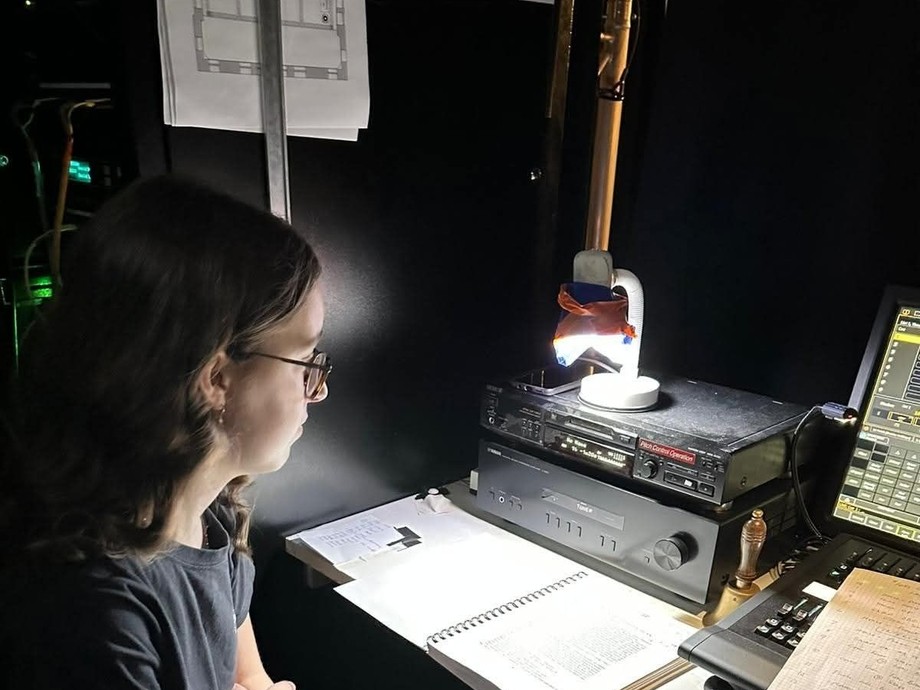 Dulcie sitting in a dimly lit room with headphones on looking at a lit up creative system (a feeling of working behind the scenes)