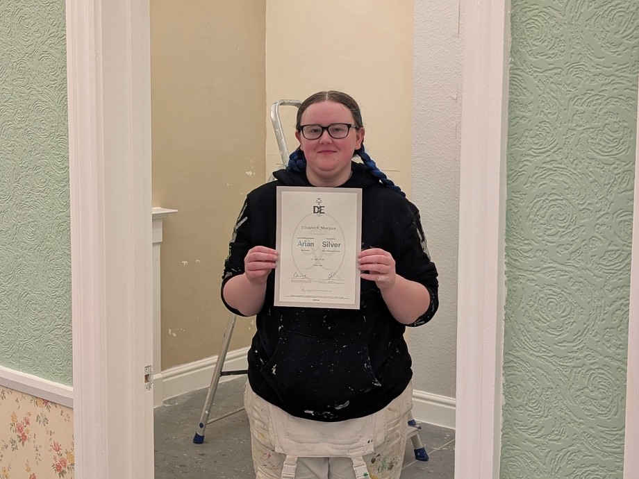 Bethan standing in a painting and decorating background holding her certificate at college