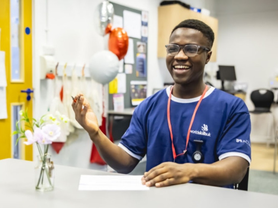 A young person in a blue t-shirt branded WorldSkillsUK at a desk wearing a lanyard smiling