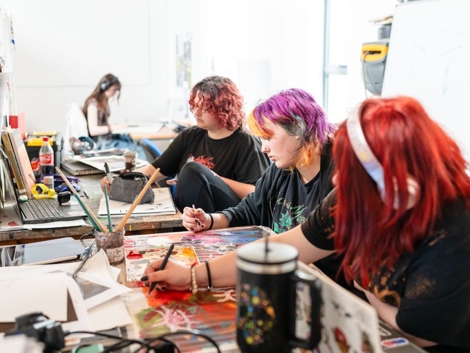 Three students with brightly coloured hair painting whilst surrounded by art supplies.