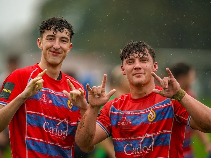 Two students in muddy rugby kit smiling at the camera and making positive hand gestures.