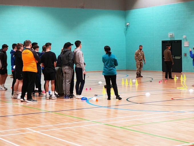 A group of students gathers around a basketball court, engaging with British Army members during an obstacle course event.