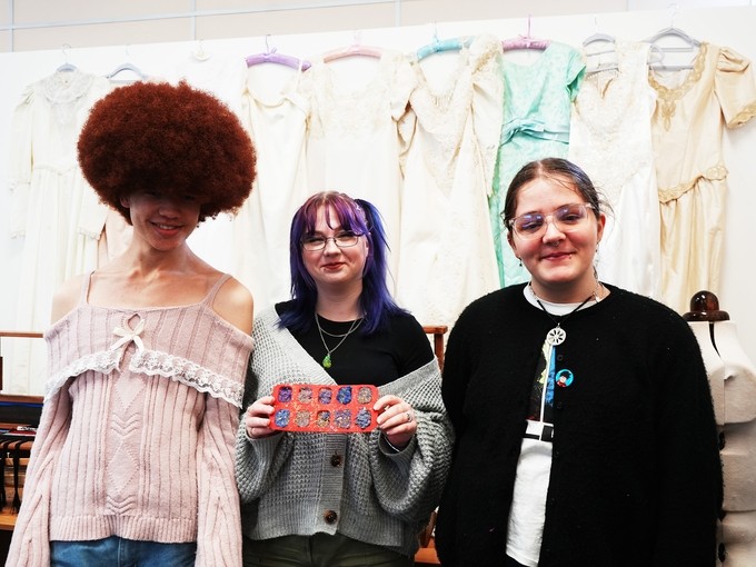Three art students with garments hanging behind them standing with items they have made